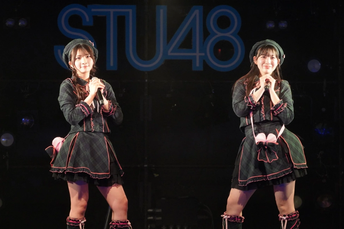 STU48のメンバーと思われる2人の女性アイドルが、チェック柄の衣装とベレー帽を着用し、マイクを持ってステージ上で笑顔でポーズをとっています。