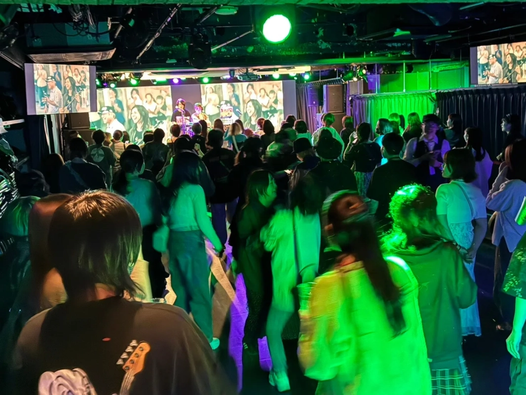 緑と紫の照明が印象的なライブハウスやクラブで、大勢の観客がステージ上のパフォーマーが映るスクリーンを見ながら盛り上がっています。活気ある雰囲気です。