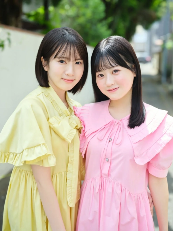 黄色のワンピースを着た女性とピンクのワンピースを着た女性の2人が、屋外で笑顔で並んで立っているポートレート写真