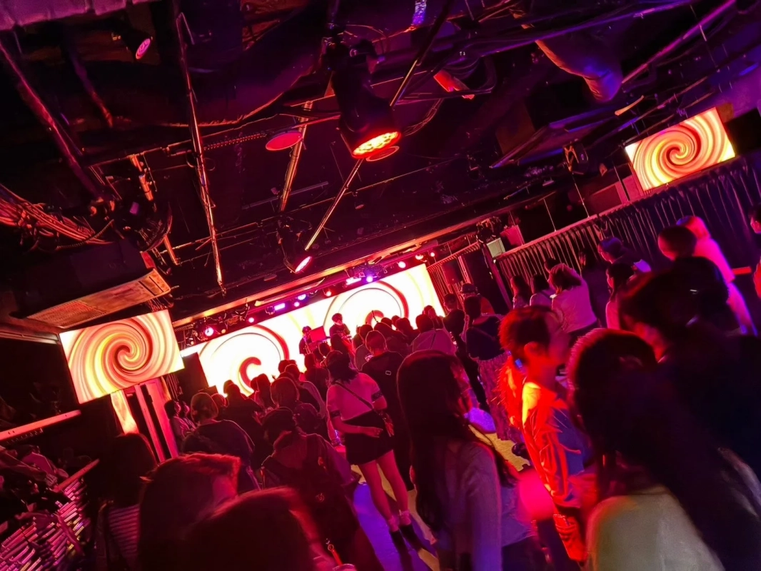 ライブハウスやクラブのような場所で、多くの観客がステージを向いている様子を捉えた画像です。赤や紫の照明が会場を照らし、ステージ背後のスクリーンには渦巻く模様が映し出されており、活気に満ちたイベントの雰囲気が伝わってきます。