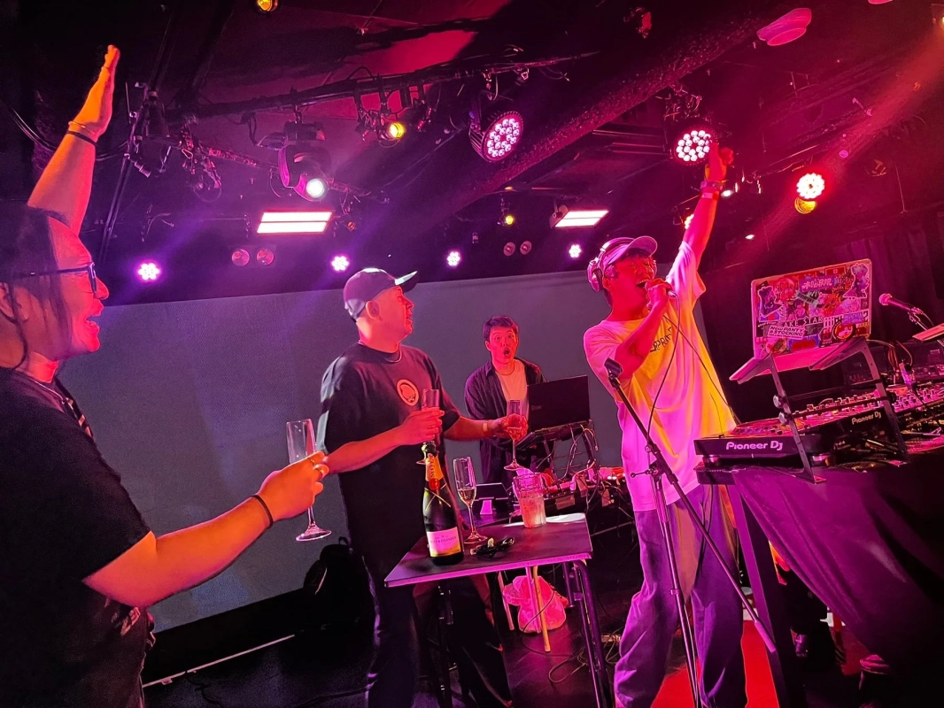 ピンクと紫の照明が照らすステージで、数人の男性がDJプレイやマイクパフォーマンスを行っています。シャンパンを手に祝う人もおり、活気ある音楽イベントの様子を捉えています。