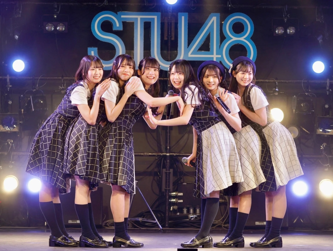 ライブステージで、チェック柄の衣装を着たSTU48のメンバー6人が笑顔でポーズをとっています。マイクを持ち、楽しそうにパフォーマンスしている様子です。