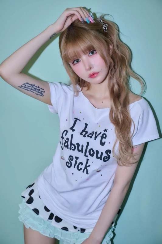 金髪の若い女性がライトブルーの背景でポーズをとっている。特徴的なメイクと派手なネイルアート、腕のタトゥーが目を引く。白Tシャツと水玉スカートを着用し、ファッション性の高いポートレート写真。