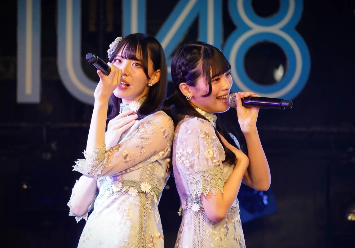 ライブステージで、マイクを手に歌う二人の女性アイドル。お揃いの刺繍入りドレスを着用し、背中合わせでパフォーマンスしている。