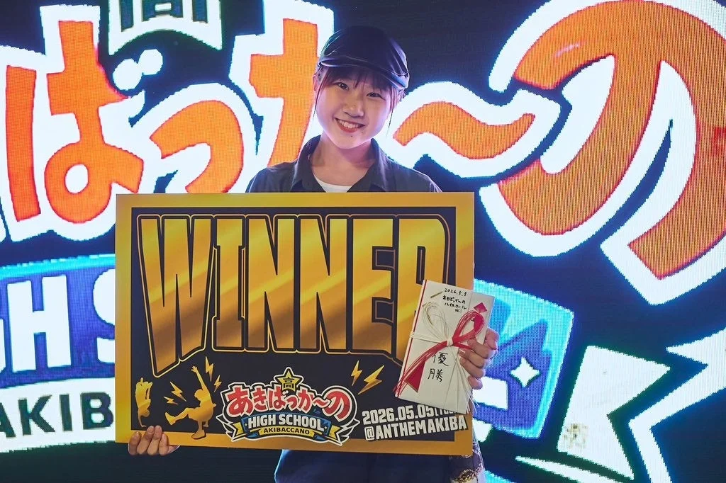 「あきばっかーのハイスクール」の大会で優勝した若い女性が、笑顔で「WINNER」のボードと賞状を掲げています。背景にはイベントロゴが映し出されたLEDスクリーンがあり、勝利の喜びを表現しています。