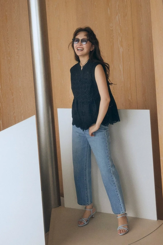 サングラスをかけ、黒いノースリーブトップスとデニムを着用した女性が、屋内で明るく笑顔を見せているポートレート