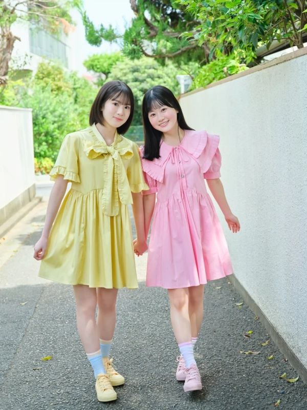 黄色とピンクのワンピースを着た二人の若い女性が、手をつないで笑顔で歩いている様子を捉えた写真