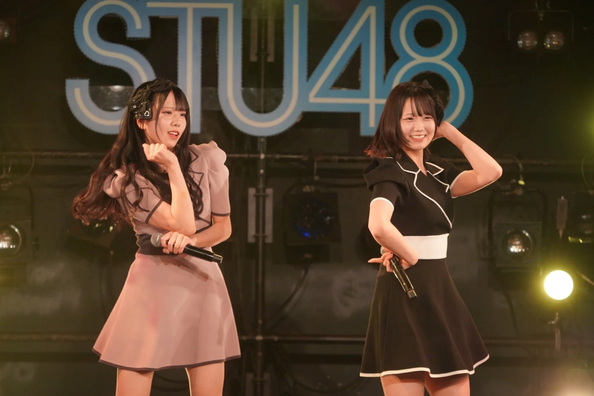 ステージ上でパフォーマンスするSTU48のメンバー2人を捉えた画像です。明るい衣装を身につけ、マイクを手に笑顔で歌っている様子が伺えます。背景にはグループ名のロゴが見えます。