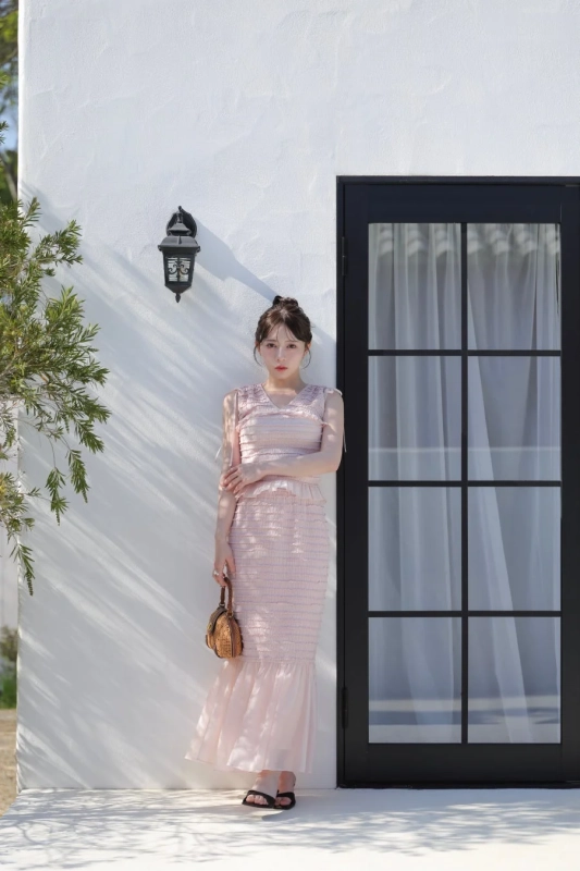 白い壁と黒いドアを背景に、ピンクのロングドレスを着た女性が立っています