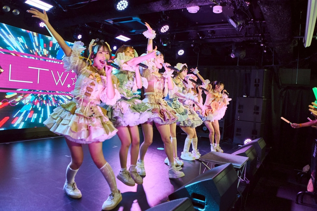 華やかな衣装をまとった女性アイドルグループが、ステージ上でライブパフォーマンスを行っている様子です。メンバーはマイクを持ち歌い、背景のLEDスクリーンには「LTW」の文字が表示され、カラフルな照明が会場を盛り上げています。