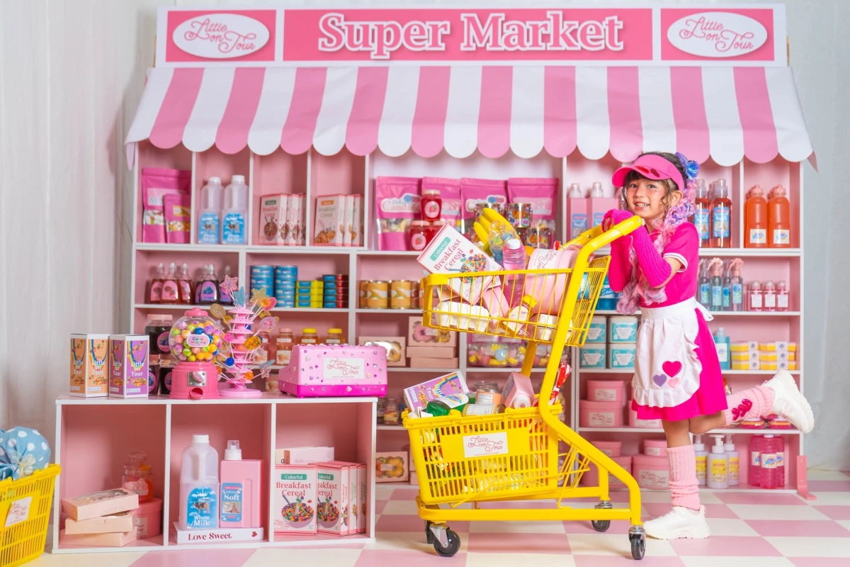 ピンクと白の可愛らしいスーパーマーケットで、ピンクの衣装の少女が黄色いショッピングカートを押している。カートには商品が満載で、少女は楽しそうな笑顔。店内には「Super Market」の看板とパステルカラーの商品が並ぶ。