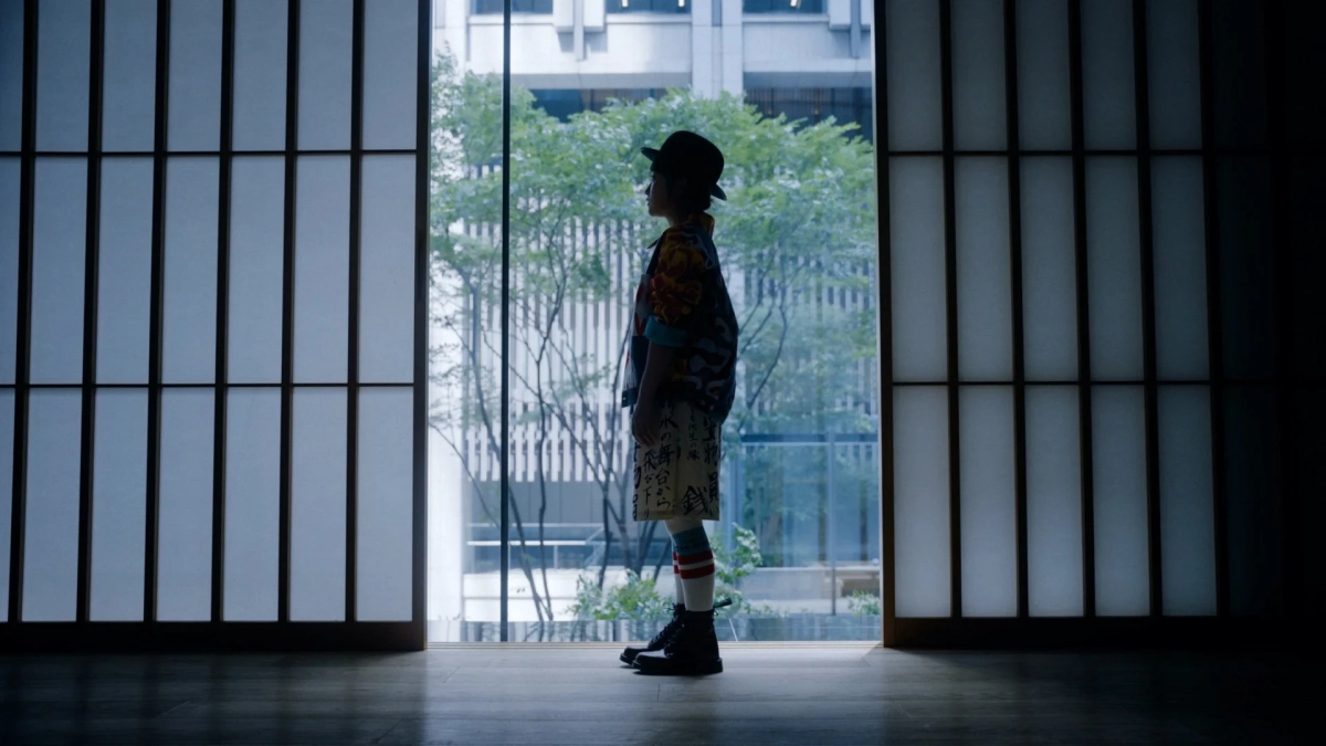 帽子をかぶった人物が、障子風の窓の前に立ち、外の緑とビルを背景にシルエットになっています。和と洋が融合したような独特のファッションが特徴的です。