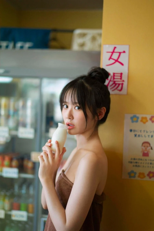 茶色いタオルを巻いた若い女性が、湯上がりに牛乳瓶の牛乳を飲んでいる。後ろには「女湯」の看板が見え、銭湯や温泉のような場所でのリラックスした様子が伺える。