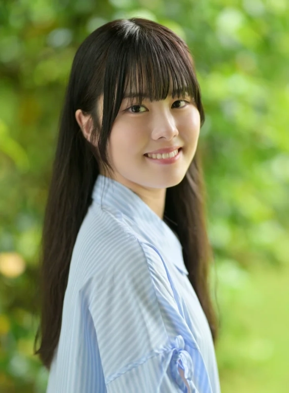 緑豊かな背景を背に、水色のストライプシャツを着た若い女性がカメラに向かって優しく微笑んでいます。