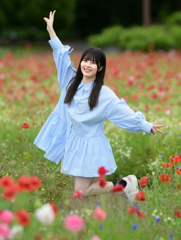 赤いポピーが咲く花畑で、青いワンピースを着た若い女性が笑顔で両腕を広げ、楽しそうにポーズをとっています。