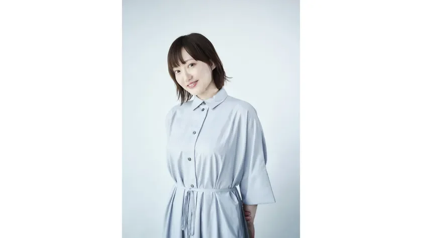 水色のシャツワンピースを着た若い女性が、白い背景の前で笑顔を見せているポートレート写真です。