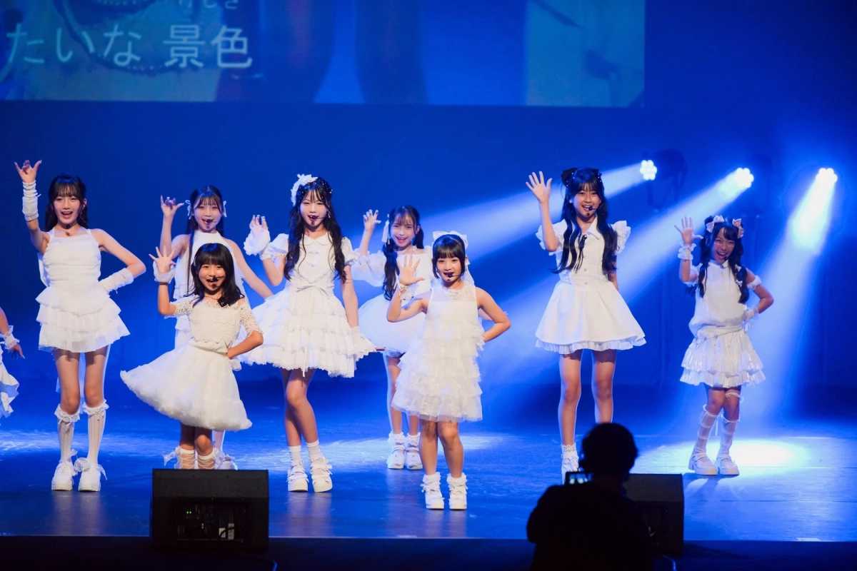 青い照明のステージで、白いフリル衣装を着た複数の少女たちが笑顔で手を振りながらパフォーマンスしている様子です。
