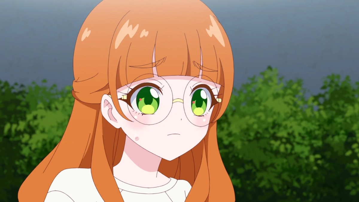 オレンジ色の髪と丸眼鏡が特徴的なアニメの少女が、屋外で少し真剣な表情を浮かべている様子。背景には緑豊かな木々が見えます。
