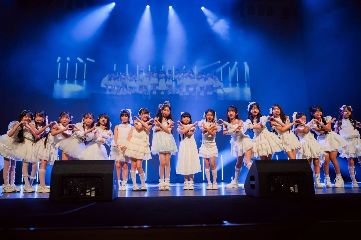 青い照明が当たるステージ上で、白い衣装を着た多くの若い女の子たちが笑顔でパフォーマンスしている様子です。