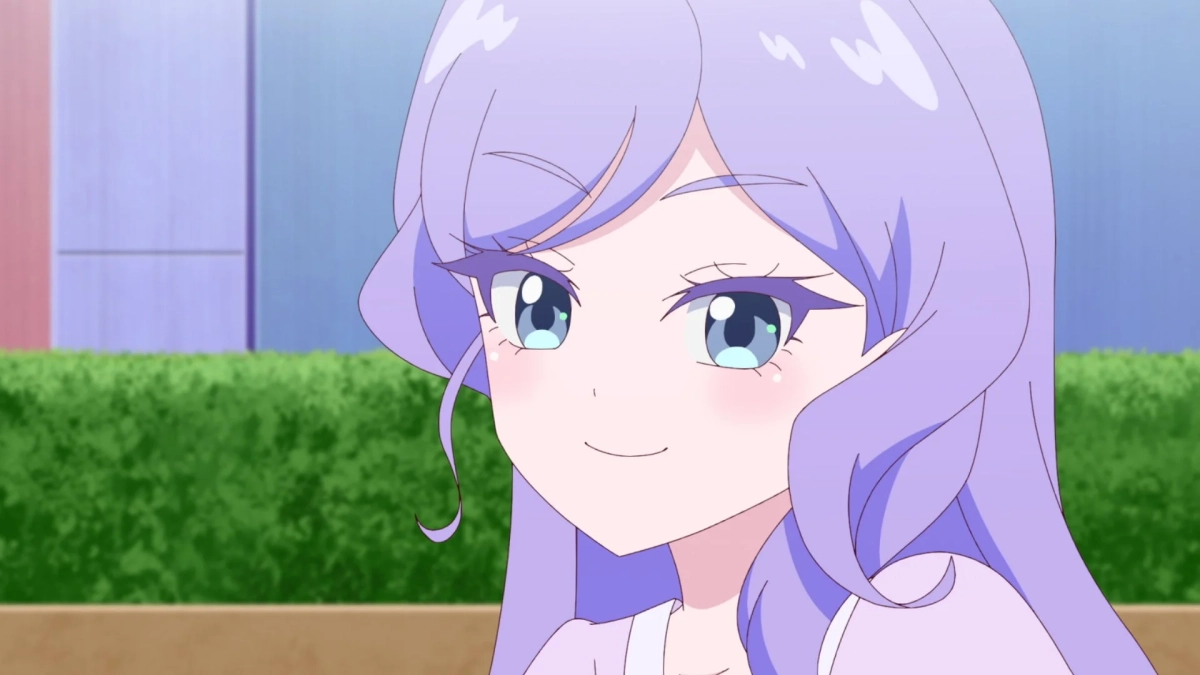 紫色の長い髪と青い目を持つアニメの女の子が、優しく微笑んでいる上半身のクローズアップ画像です。背景はぼかされており、緑の植え込みが見えます。