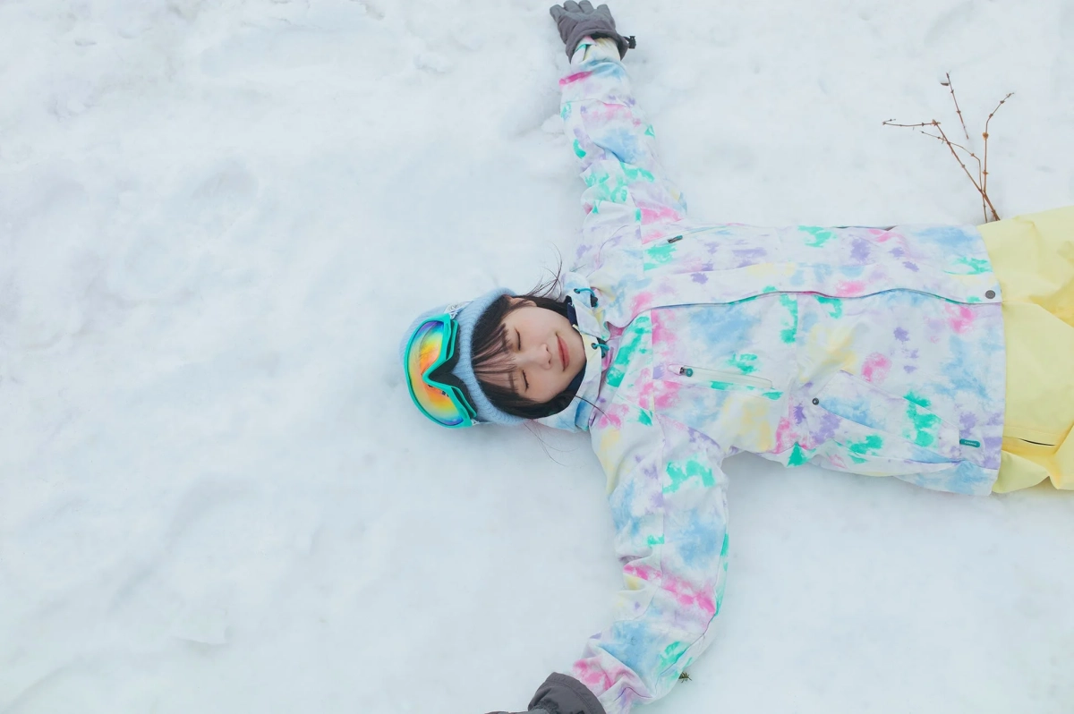 カラフルなスキーウェアを着用した若い女性が、雪の上に仰向けに寝転がり、目を閉じてリラックスした表情を浮かべています。