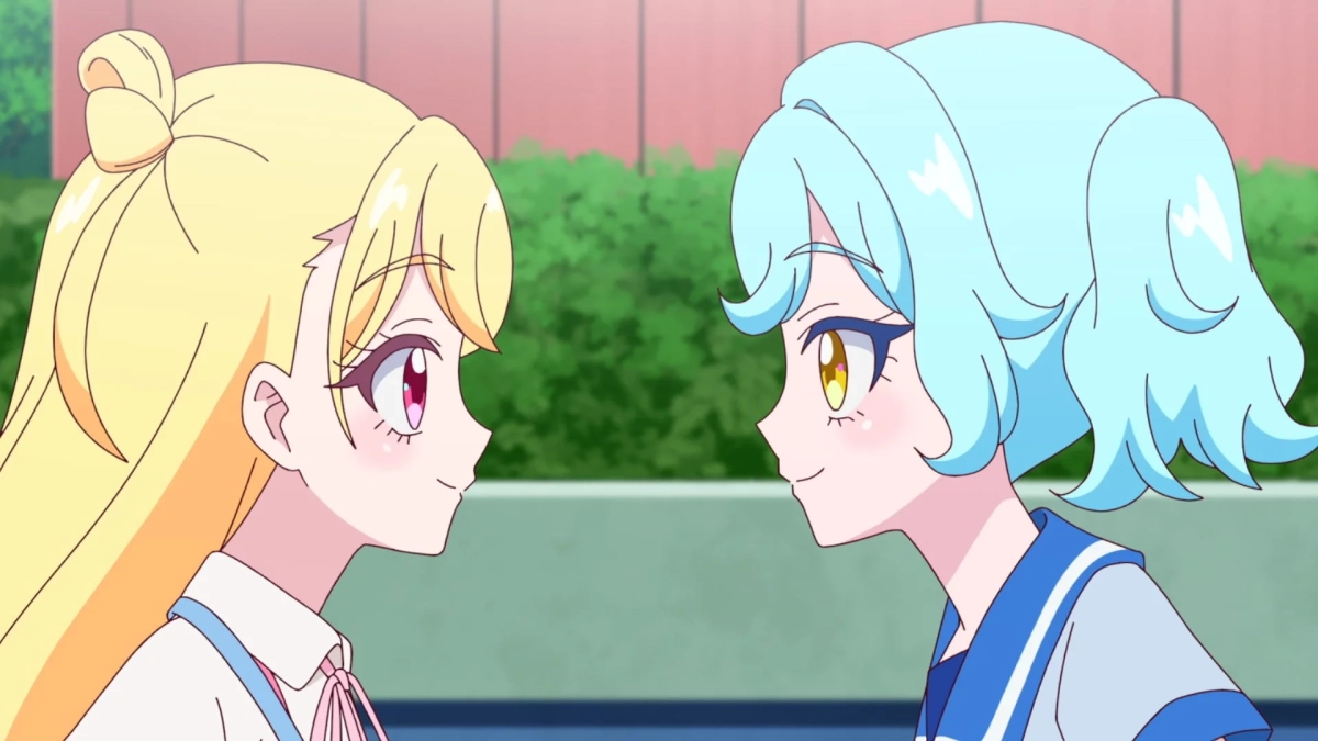 アニメ風のイラストで、二人の少女が向かい合って微笑んでいます。一人は長い金髪にピンクの瞳、もう一人は水色の短い髪に黄色の瞳です。二人とも制服を着用しており、親しげな様子が描かれています。