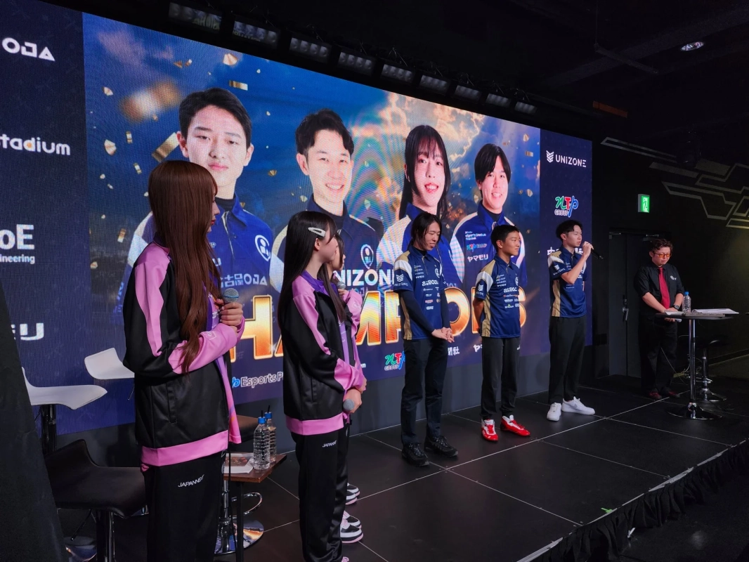 eスポーツイベントの様子で、複数の人々がステージ上に立ち、後方の大型スクリーンにはeスポーツ選手らしき人物の写真が映し出されています。