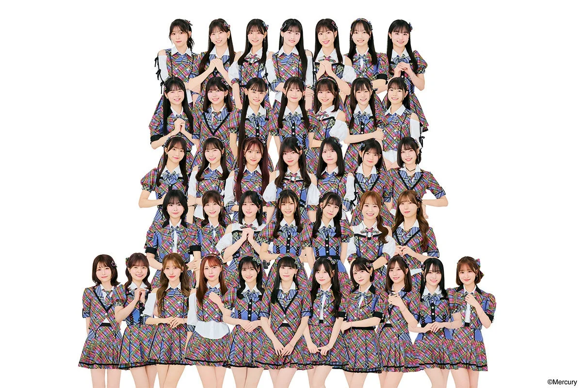 白い背景に、カラフルなチェック柄の衣装をまとった多数の女性アイドルグループが、笑顔でポーズをとる集合写真です。