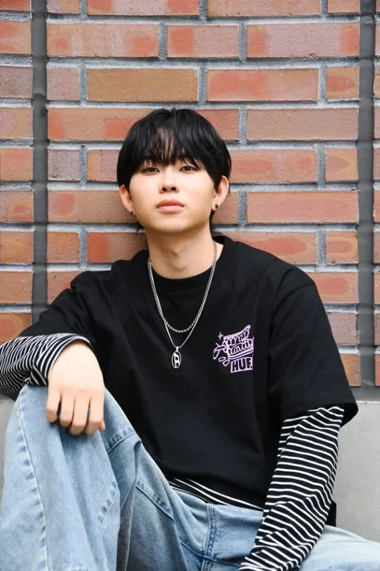 レンガの壁を背景に座る若い男性のポートレートです。黒いレイヤードTシャツと水色のデニムを着用し、銀色のネックレスをしています。