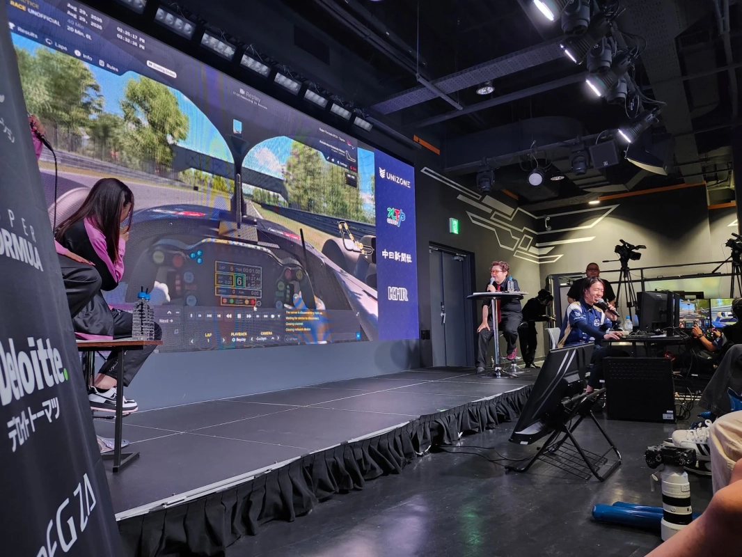 eスポーツイベントで、大型スクリーンにレーシングゲームが映し出され、ステージ上では実況者がマイクを使って解説しています。