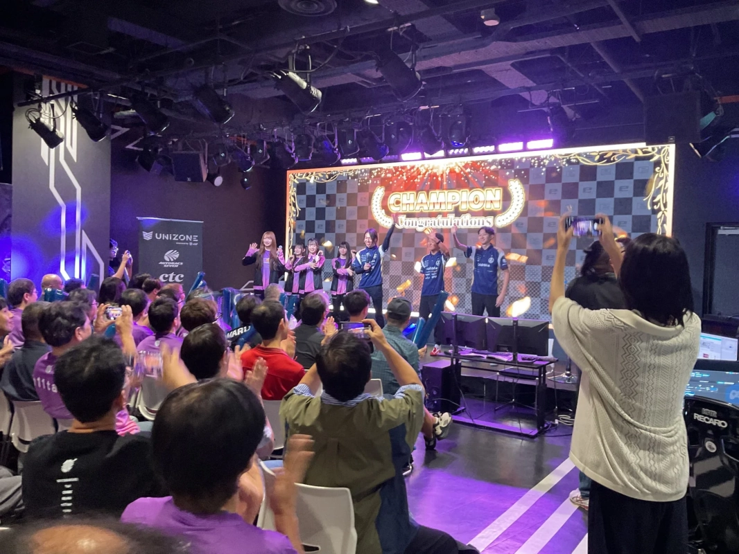 eスポーツイベントの表彰式で、ステージ上のチャンピオンと出演者が観客に手を振っている様子。