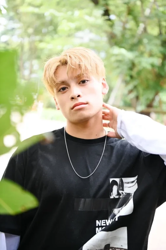 金髪の若い男性が屋外でカメラ目線でポーズをとっているポートレート写真です。黒いTシャツとシルバーのネックレスを着用し、背景には緑豊かな木々がぼやけて写っています。