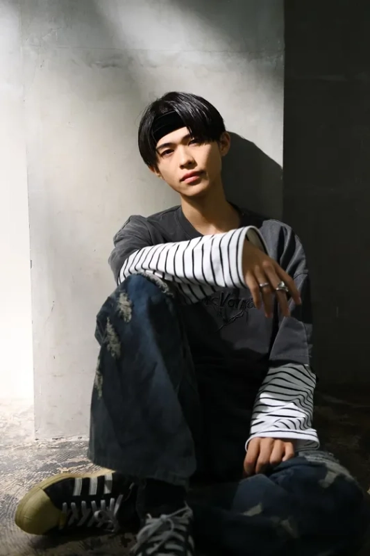 黒いヘッドバンドとダメージジーンズを着用した若い男性が、壁にもたれて座っているポートレート。ストリート系のファッションで、やや影のある雰囲気の屋内ショットです。